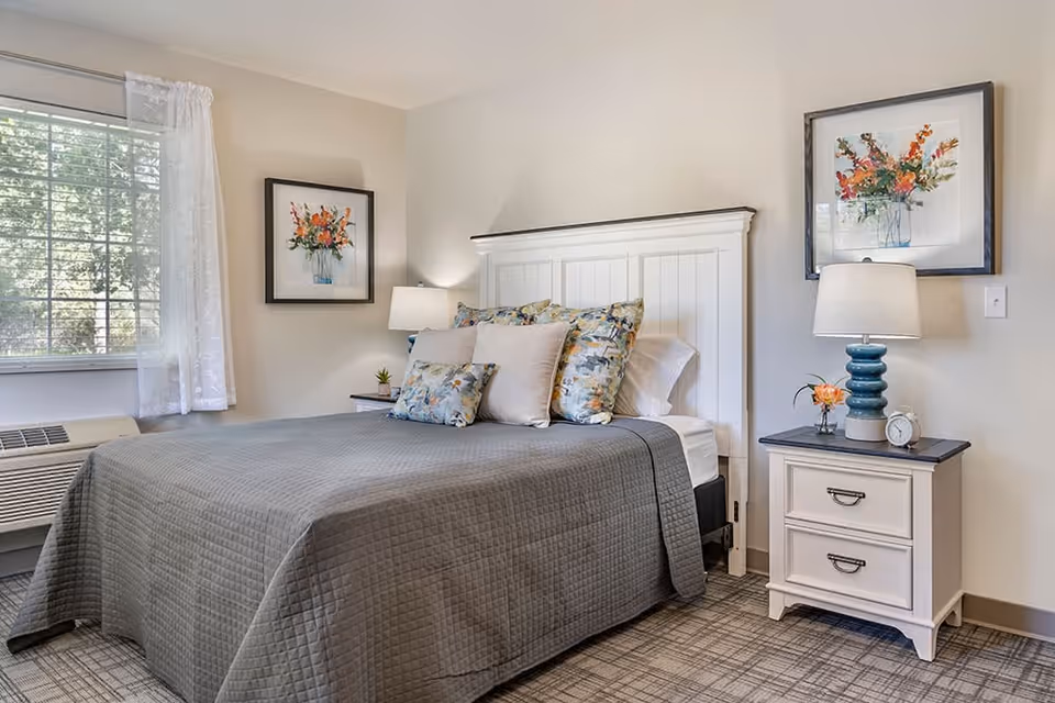 A neatly made bed with a gray quilt and multiple decorative pillows in a bedroom. The room has a white headboard, two framed floral artworks on the walls, a nightstand with a blue lamp, a small flower vase, and an alarm clock. A window with white curtains allows natural light to enter the room.