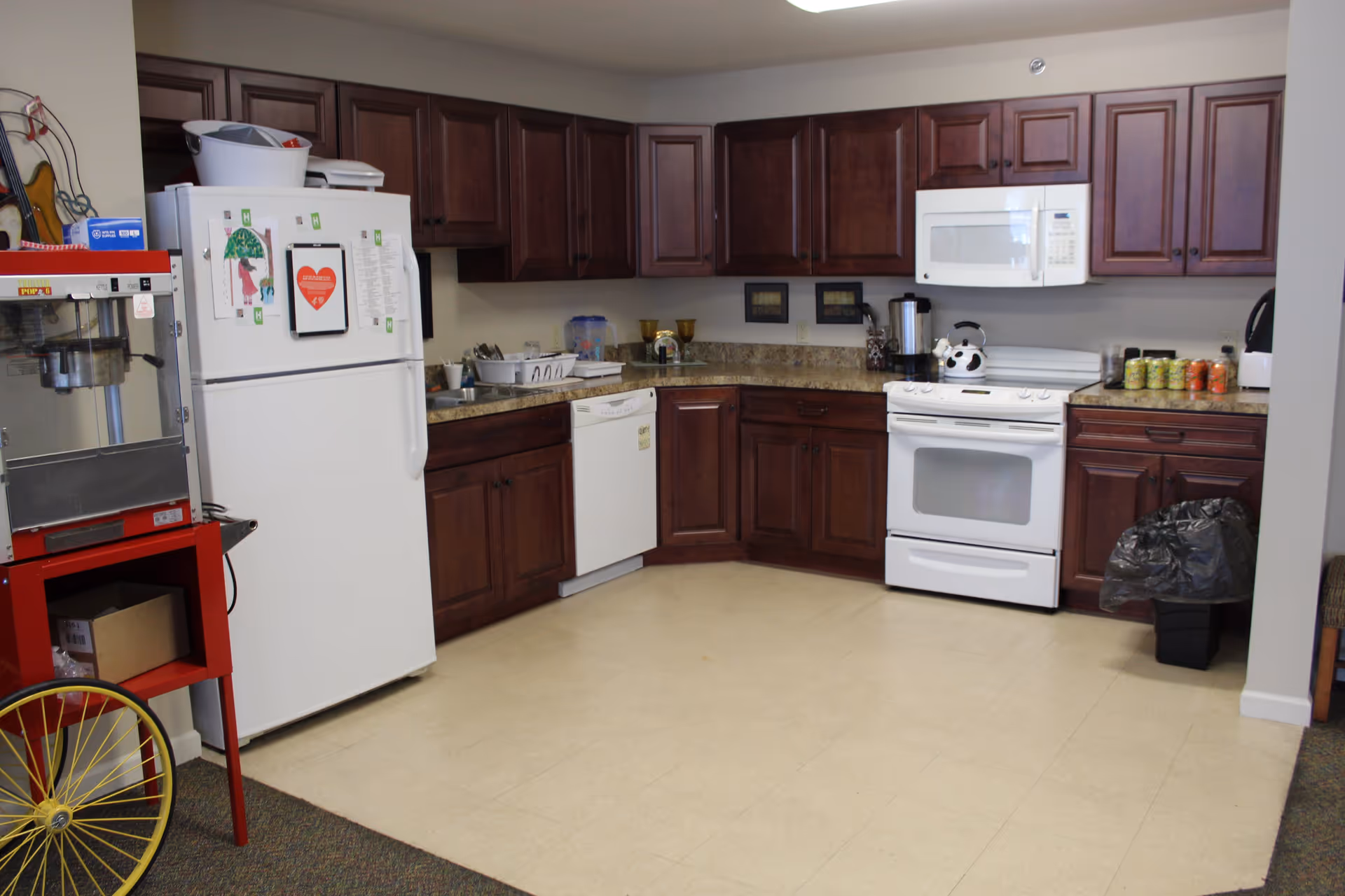 A kitchen area with dark wooden cabinets, a white refrigerator with magnets and papers on it, a white dishwasher, a white stove with a kettle on top, a white microwave above the stove, and a red popcorn machine on the left side. The floor is light-colored linoleum and there is a trash bin with a black bag on the right side.