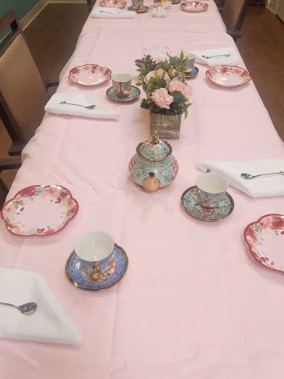 A long table covered with a pink tablecloth set for tea with teacups, saucers, floral plates, napkins, spoons, a decorative teapot and a floral centerpiece.