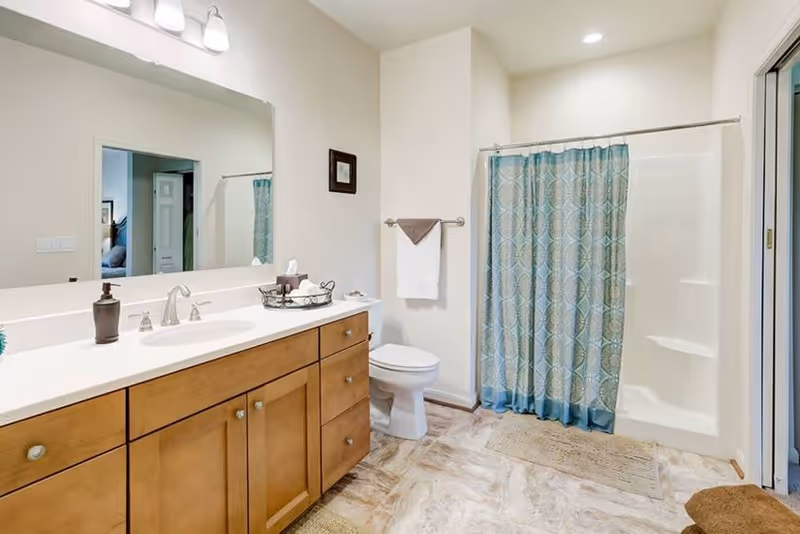 A clean and bright bathroom featuring a wooden vanity with a white countertop and sink, a large mirror above the vanity, a toilet, and a shower with a blue patterned curtain. The floor has a light-colored tile, and there are towels hanging on a rack next to the toilet.