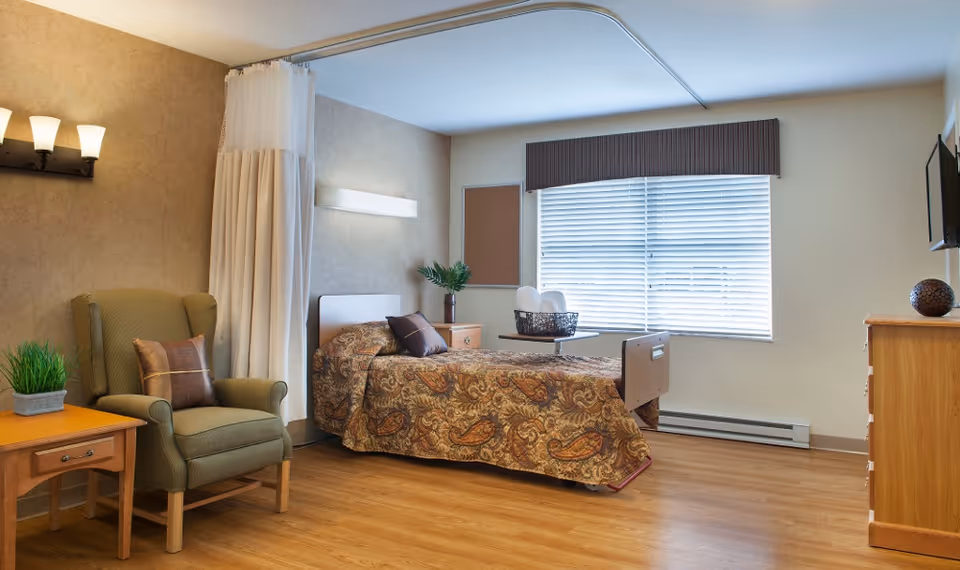 A well-lit senior living bedroom featuring a single bed with a patterned bedspread, a green armchair with a pillow, a wooden side table with a plant, a window with blinds, a wall-mounted TV, and a wooden dresser. The room has light-colored walls and wood flooring.
