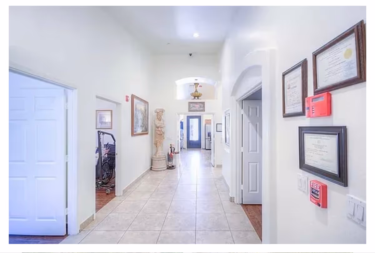 Bright tiled hallway in a senior living facility with doors on either side, framed certificates and fire alarms on the right wall, and a statue near the far end.