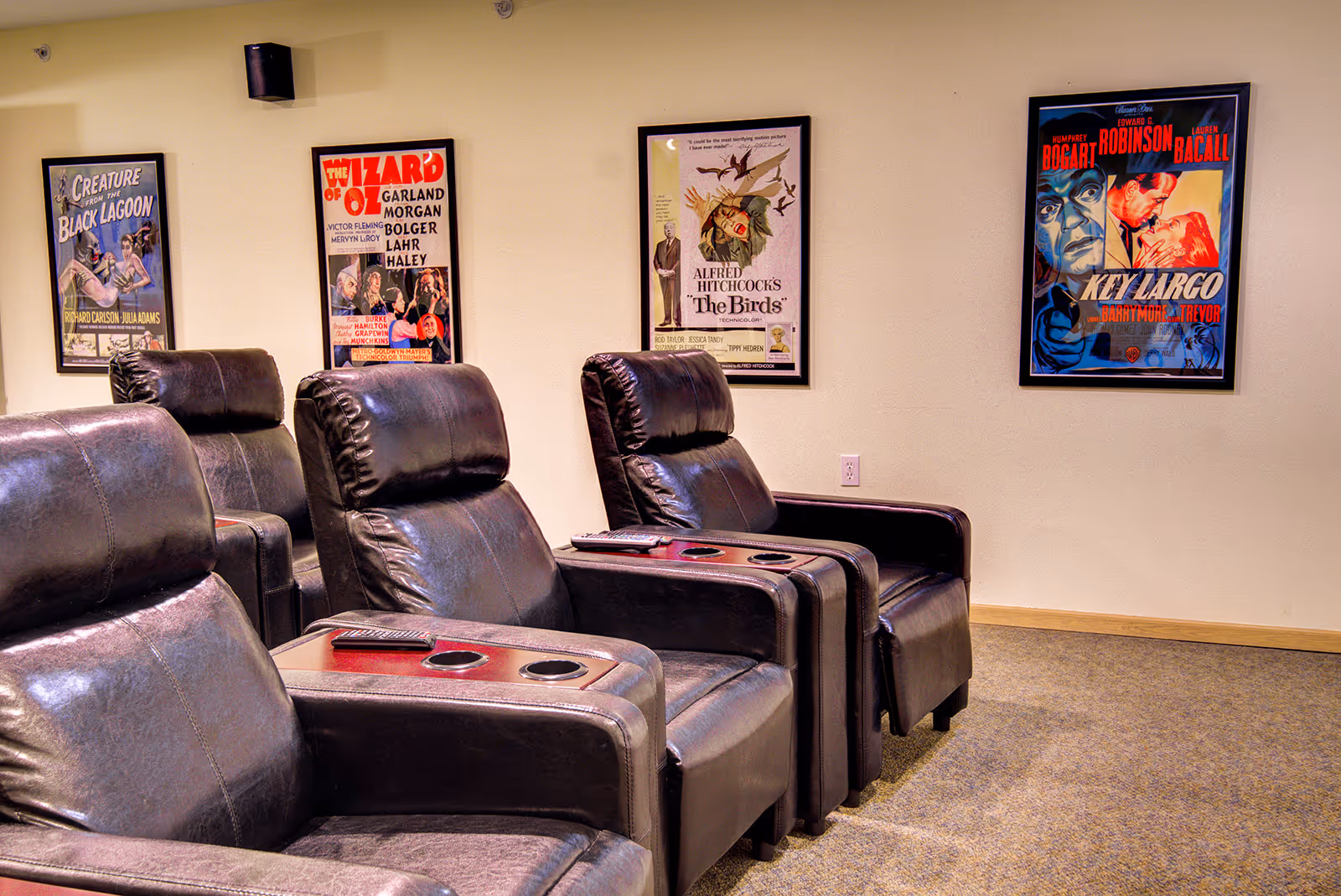 A cozy movie theater room with four black leather recliner chairs arranged in two rows. Each chair has cup holders and a remote control on the armrest. The beige wall behind the chairs is decorated with framed vintage movie posters including Creature from the Black Lagoon, The Wizard of Oz, Alfred Hitchcock's The Birds, and Key Largo.