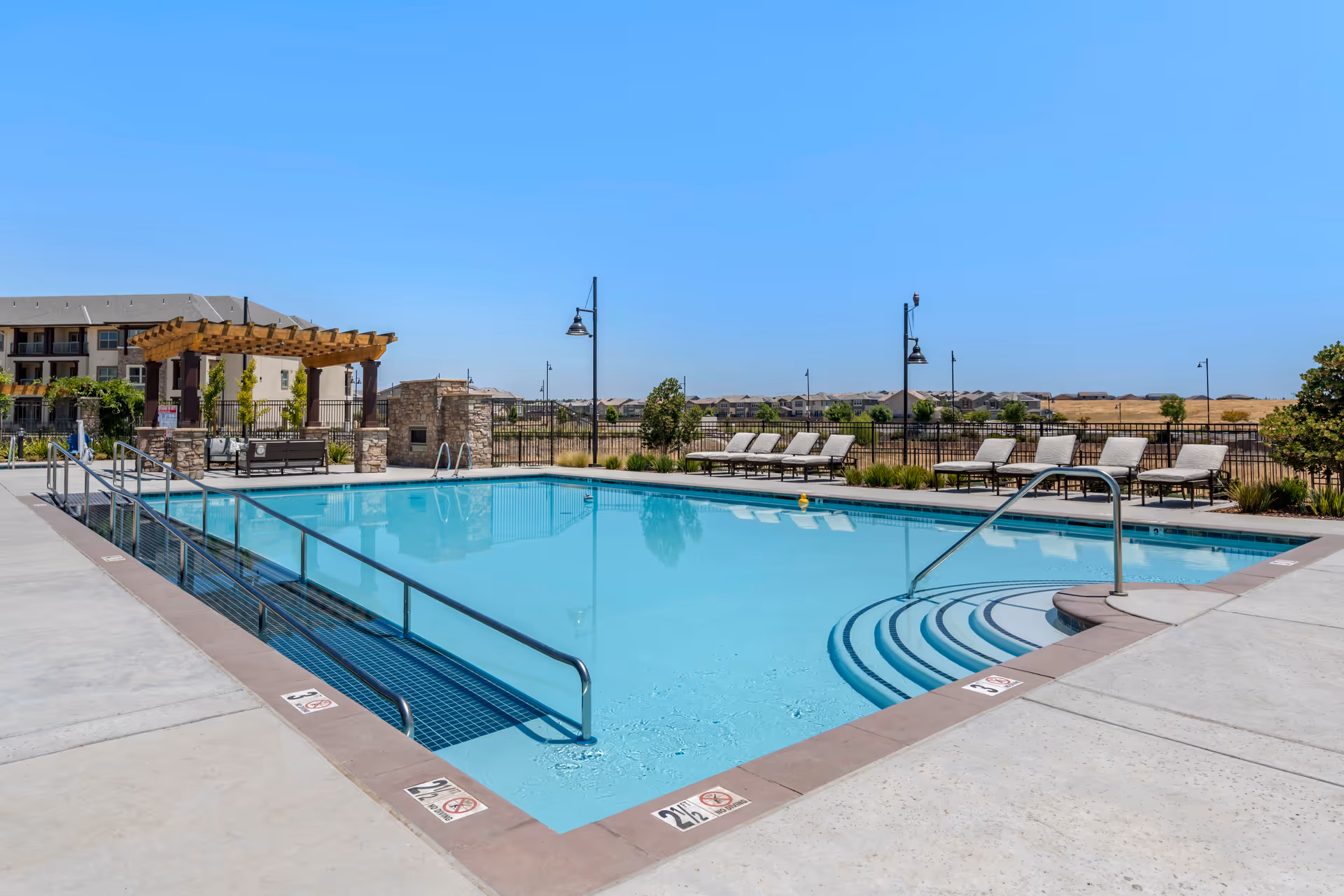 Outdoor swimming pool with lounge chairs, a pergola, and apartment buildings in the background under a clear blue sky.