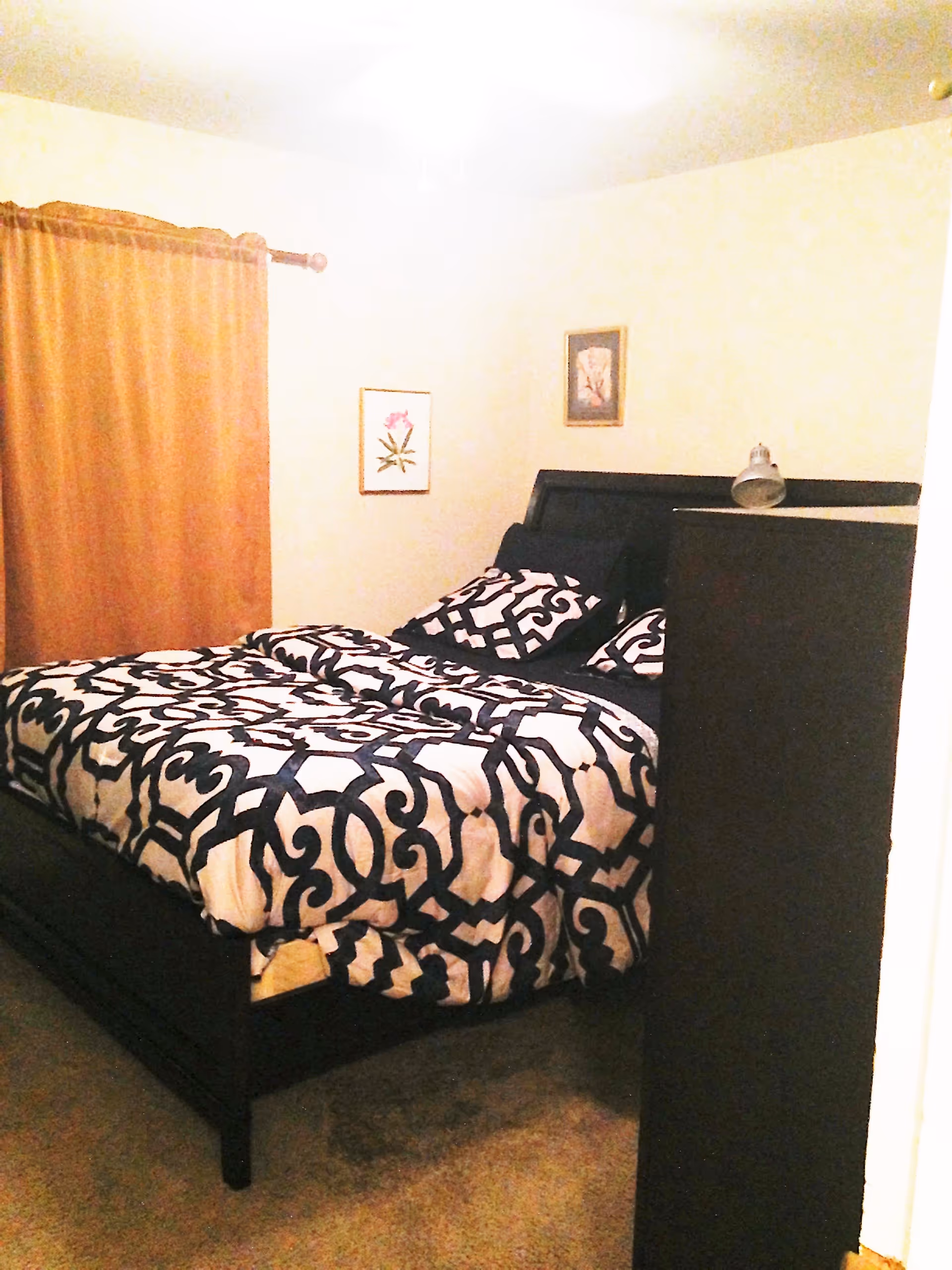 A bedroom with a bed featuring a black and white patterned comforter and pillows. There are two framed pictures on the wall above the bed and a window with brown curtains to the left. A dark-colored dresser with a small lamp on top is positioned near the foot of the bed.