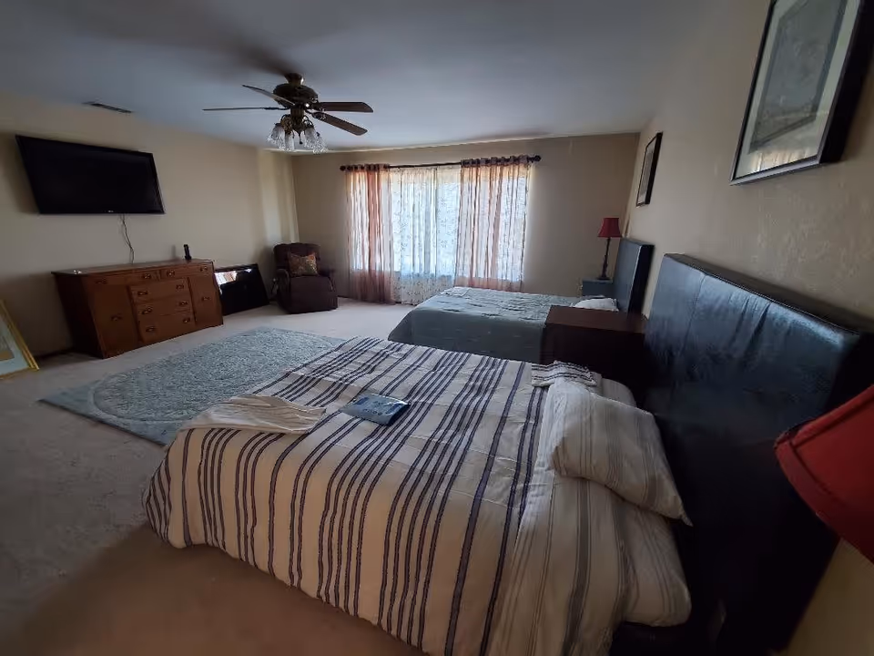 A spacious bedroom with two beds, one with striped bedding and the other with solid bedding. The room features a ceiling fan with lights, a large window with sheer and opaque curtains, a wooden dresser with a flat-screen TV mounted above it, a recliner chair in the corner, and framed artwork on the walls.