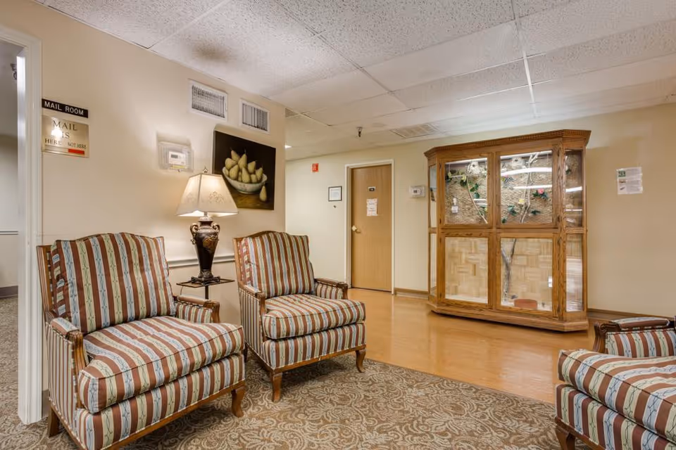 A sitting area in a senior living facility with striped armchairs, a table lamp, wall art, and a wooden display cabinet.