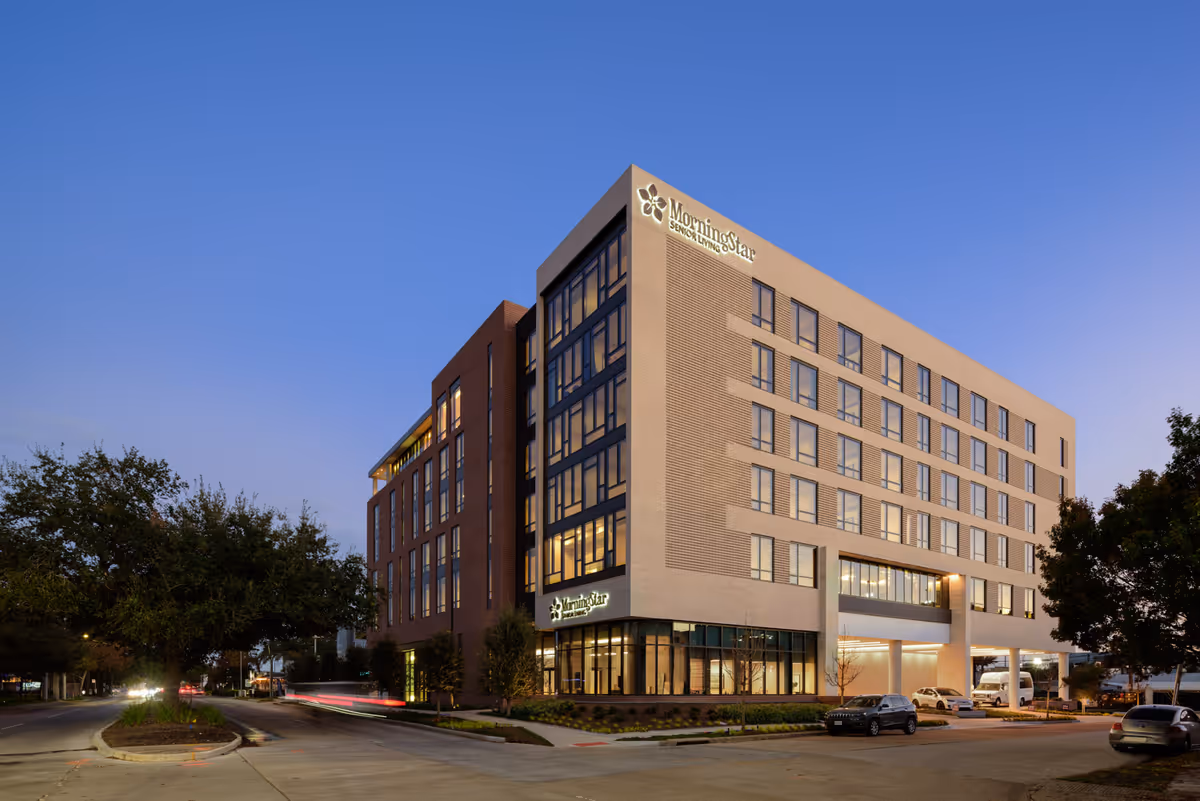 Exterior view of a modern multi-story building at dusk with the sign 'MorningStar Senior Living' visible on the corner. The building has large windows and is surrounded by trees and parked cars.