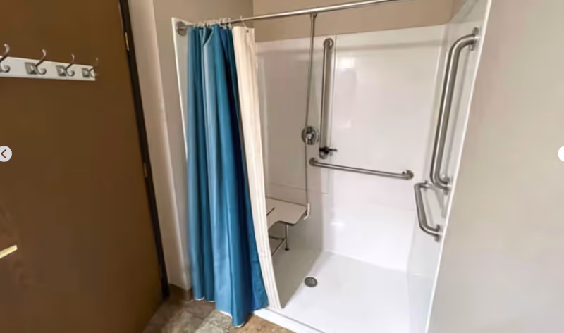 Accessible shower area with white walls, multiple stainless steel grab bars, a fold-down shower seat, and a blue and white shower curtain. A brown door with hooks is visible to the left.