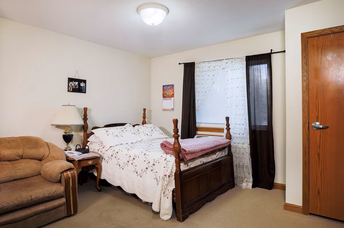 A cozy bedroom with a wooden four-poster bed covered in a white floral quilt and a folded pink blanket at the foot. Next to the bed is a wooden nightstand with a lamp and some items on it. To the left of the nightstand is a brown upholstered armchair. A window with sheer white and dark brown curtains is behind the bed. A wooden door is visible on the right side of the room.