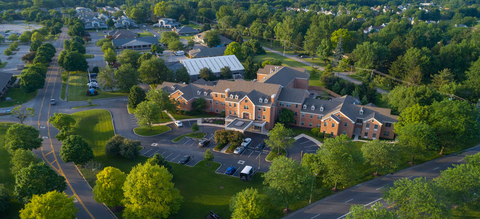 Aerial view of a senior living facility named Harmony Trace surrounded by lush green trees and landscaped grounds. The building is a large, multi-wing brick structure with a parking lot in front and roads surrounding the property. The area around the facility includes additional buildings and residential neighborhoods.