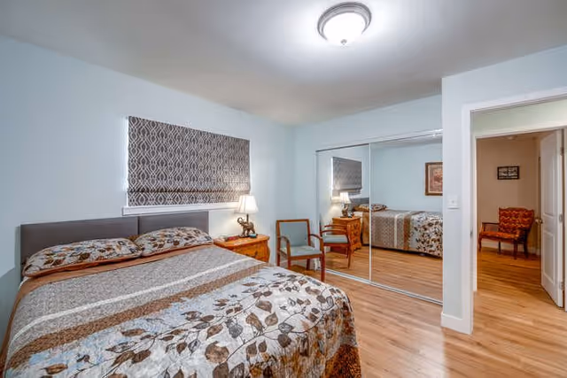 A bedroom with a bed covered in a floral patterned quilt, a wooden nightstand with a lamp, two wooden chairs, a window with a patterned blind, and a large mirrored closet door reflecting the room.