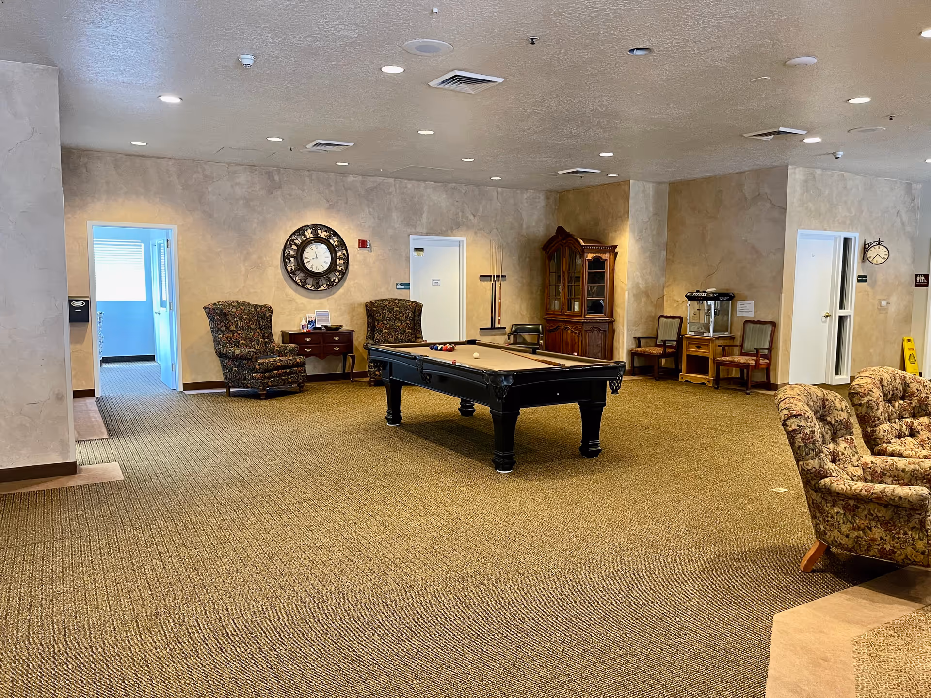 A carpeted assisted living common room featuring a central pool table, upholstered armchairs, wooden cabinets, and wall clocks.