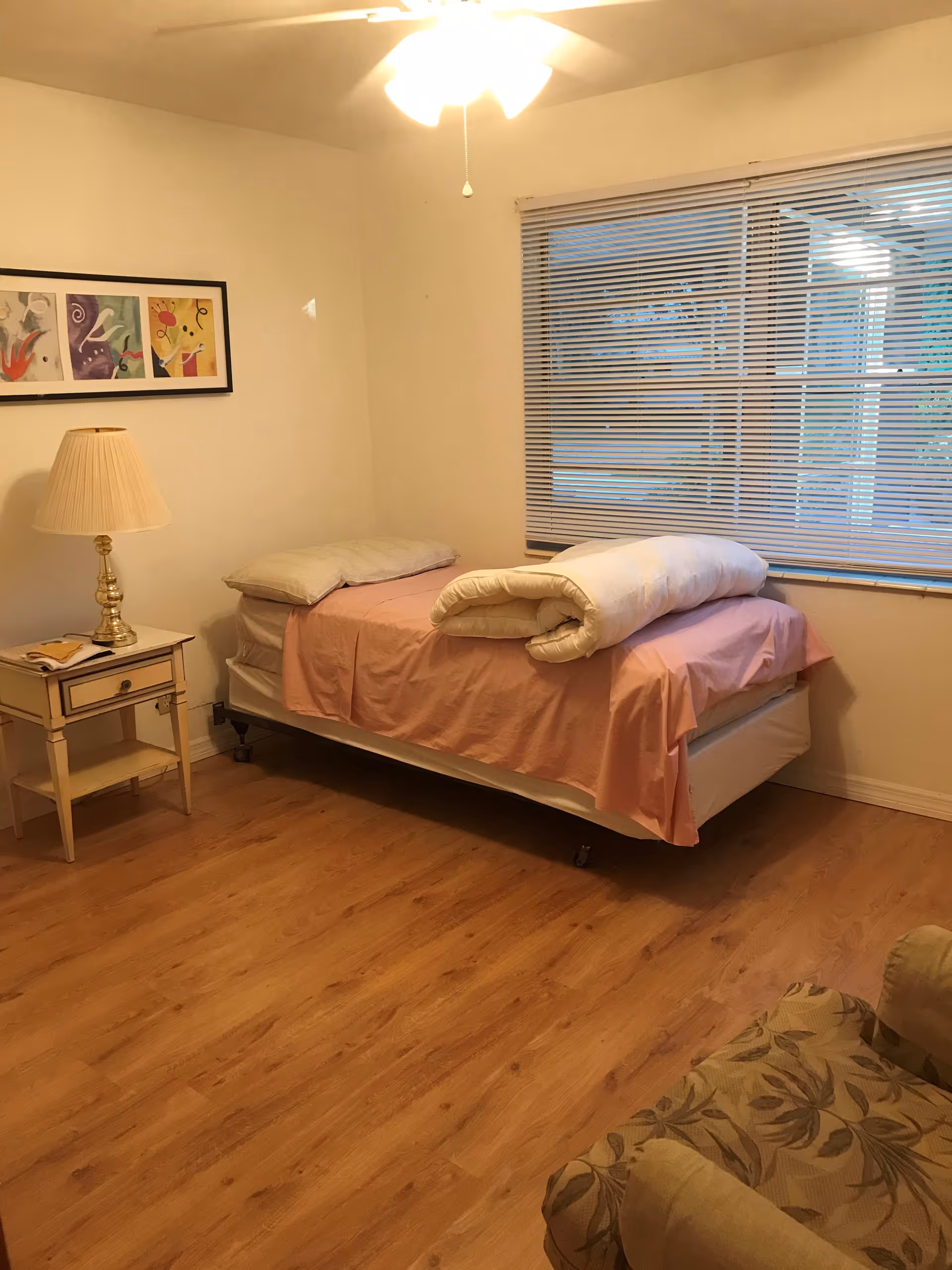 A simple bedroom with a single bed covered in pink sheets and a white comforter folded on top. There is a wooden nightstand with a lamp and a book beside the bed. A large window with closed blinds is on the right wall, and a ceiling fan with a light is on the ceiling. The floor is wooden, and a floral patterned armchair is partially visible in the foreground.