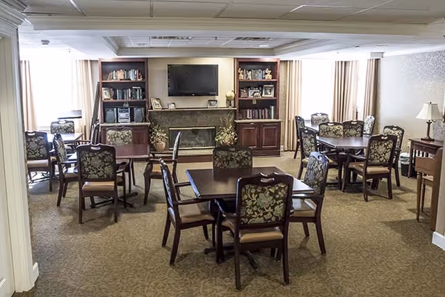 Common room with multiple dining tables and upholstered chairs facing a TV above a fireplace flanked by bookshelves.