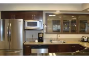 Modern kitchen area with stainless steel refrigerator, microwave, dishwasher, coffee maker, and toaster. Dark wood cabinets with glass-front upper cabinets and a double sink under a window.
