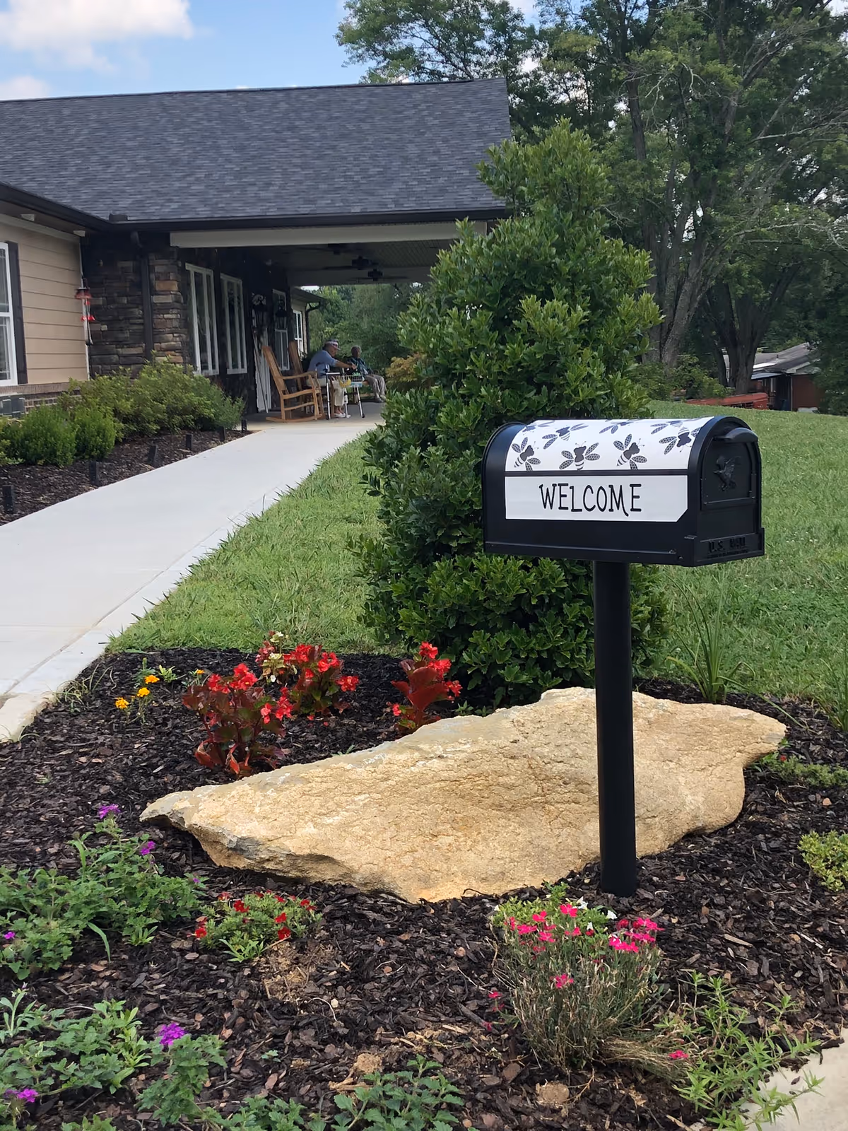 A landscaped garden area with colorful flowers and a large rock in front of a black mailbox that has a white cover with leaf patterns and the word 'WELCOME' on it. In the background, there is a building with a covered porch where two people are sitting on rocking chairs. The scene is outdoors with green grass and trees.
