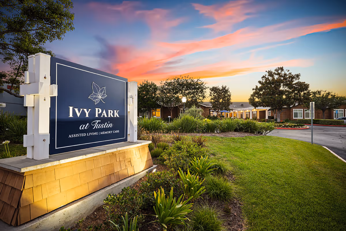 Entrance sign for Ivy Park at Tustin assisted living and memory care facility with landscaped greenery and trees, set against a colorful sunset sky.