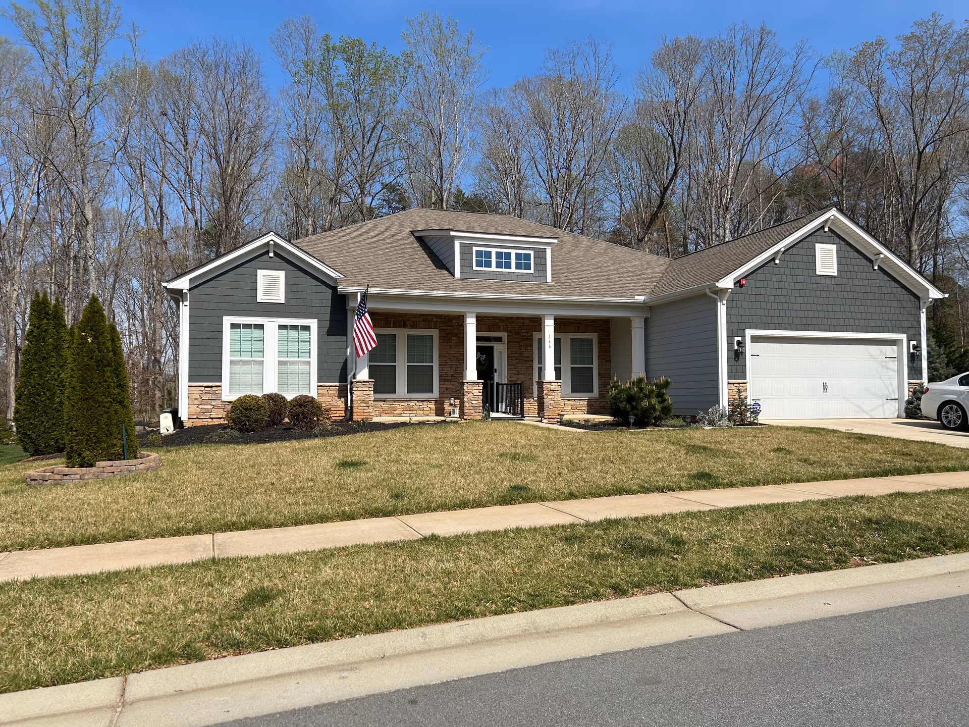 Single-story gray ranch-style house with a two-car garage, front porch displaying an American flag, and a lawn with trees behind it.