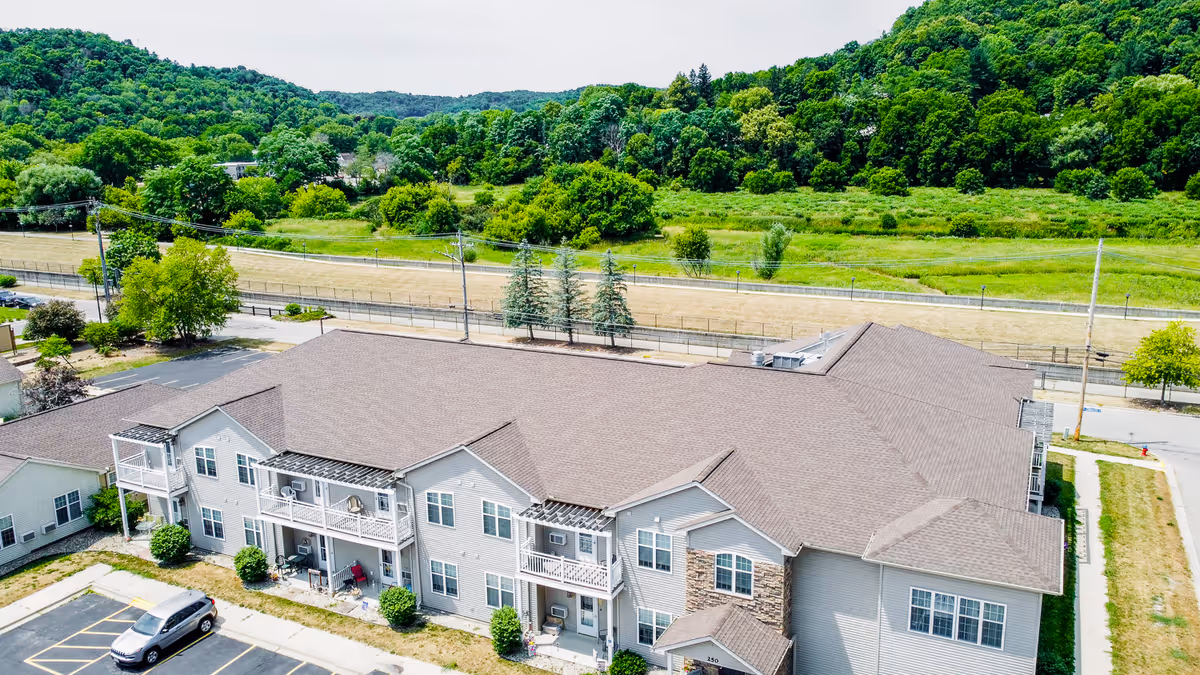 Aerial view of a two-story senior apartment building with balconies, parking spaces, and surrounding tree-lined landscape.