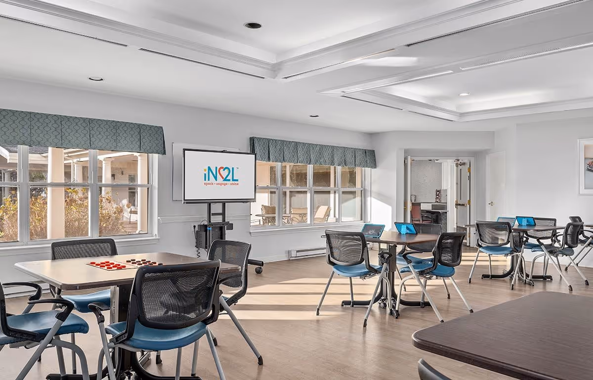 A bright and spacious activity room with several tables and chairs arranged for group activities. A large screen on a stand displays the logo 'iN2L spark engage shine'. The room has large windows with blue valances letting in natural light, and light-colored walls and flooring. Some tables have tablets or game pieces on them.