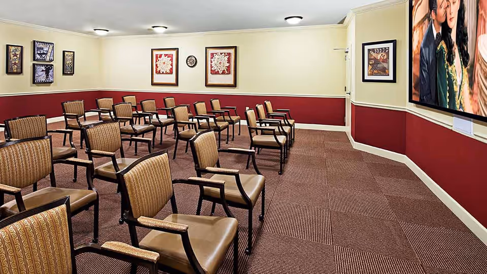 A room with multiple rows of brown cushioned chairs arranged facing a large wall-mounted television screen displaying a scene from a movie. The walls are painted cream and red, decorated with framed artwork and a clock. The floor is carpeted with a patterned design.