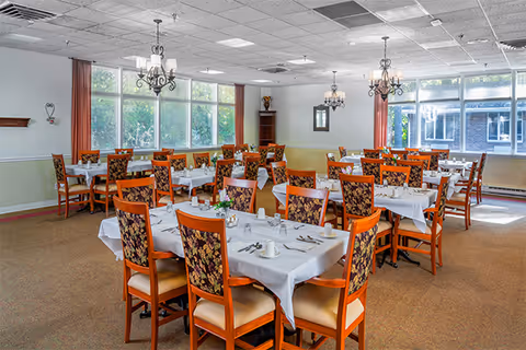 A bright dining room with multiple tables covered in white tablecloths, each set with cups, saucers, and silverware. The chairs have wooden frames with floral-patterned upholstery. Large windows let in natural light, and chandeliers hang from the ceiling. The room has beige carpeting and light-colored walls with red curtains on the windows.