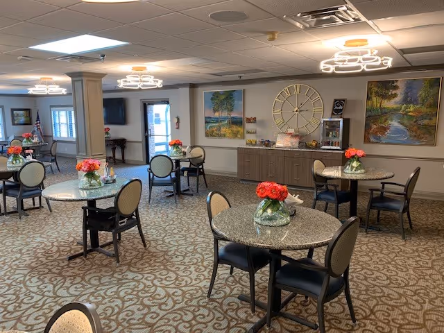 A well-lit common area with multiple round tables, each adorned with a vase of red flowers. The room has patterned carpet flooring, modern ceiling lights, and several chairs around the tables. The walls feature large paintings and a decorative clock. There is a cabinet against the wall with some items on top, and a door and windows are visible in the background.