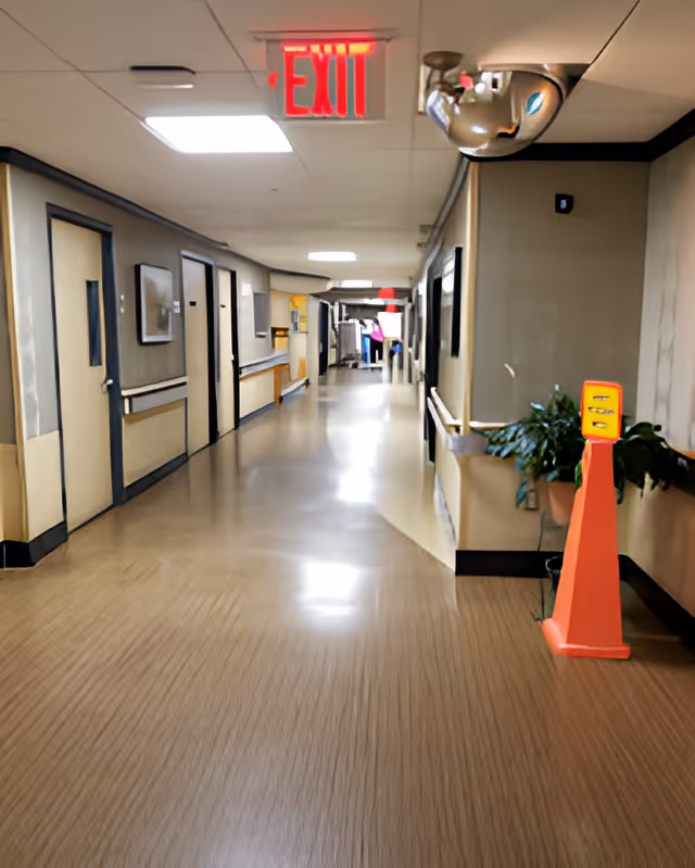 A long, clean hallway in a senior living facility with beige walls and wood-patterned flooring. Several closed doors line the left side, and handrails run along both walls. A red exit sign is illuminated on the ceiling. There is a convex security mirror mounted on the ceiling corner on the right side, and a bright orange caution cone stands near a potted plant on the right.