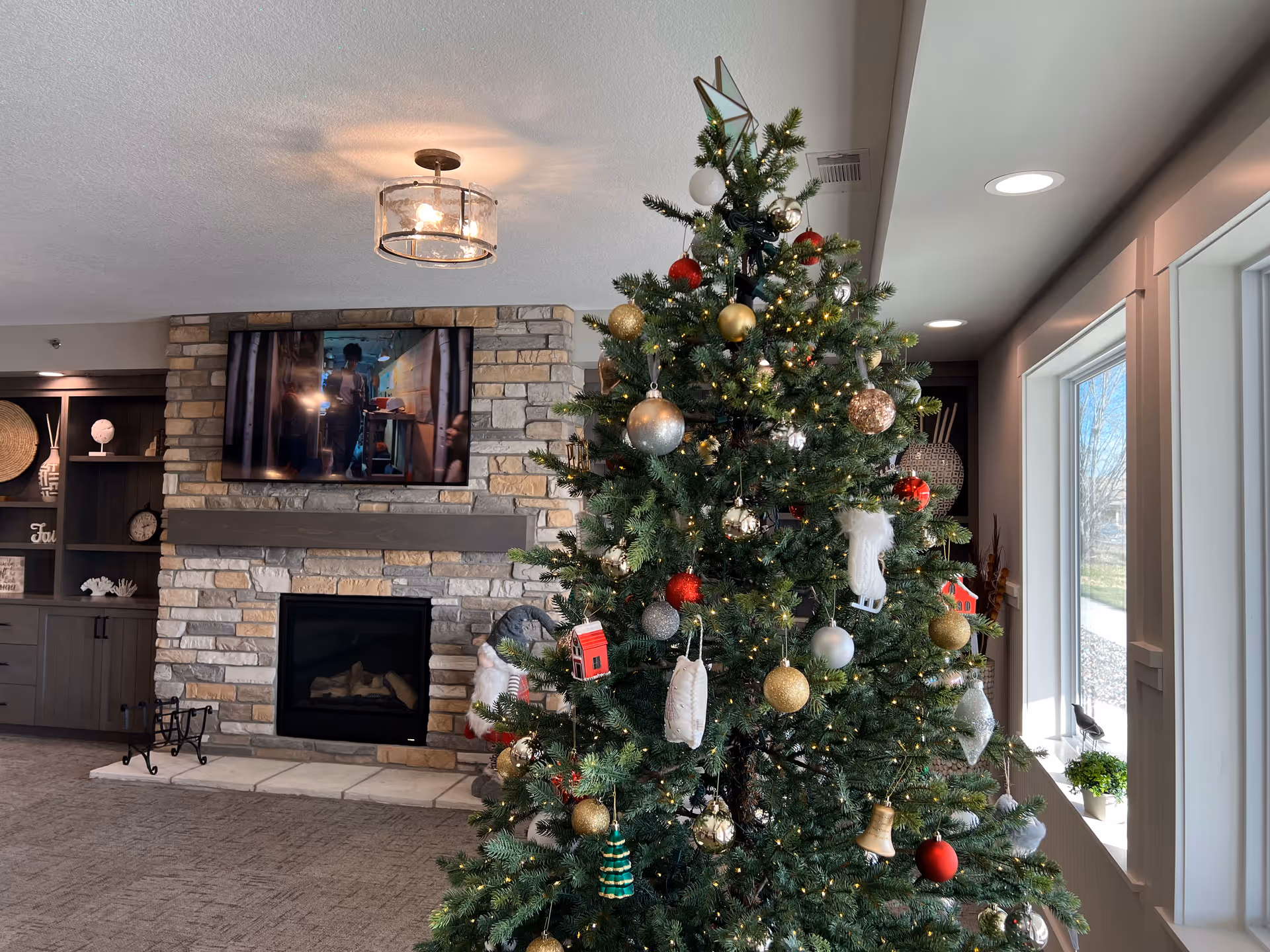 A decorated Christmas tree with various ornaments including red, gold, and silver balls, a small red house, and other festive decorations stands in a cozy living room. Behind the tree is a stone fireplace with a mounted flat-screen TV above it. To the left, there is a built-in shelving unit with decorative items. Large windows on the right let in natural light.