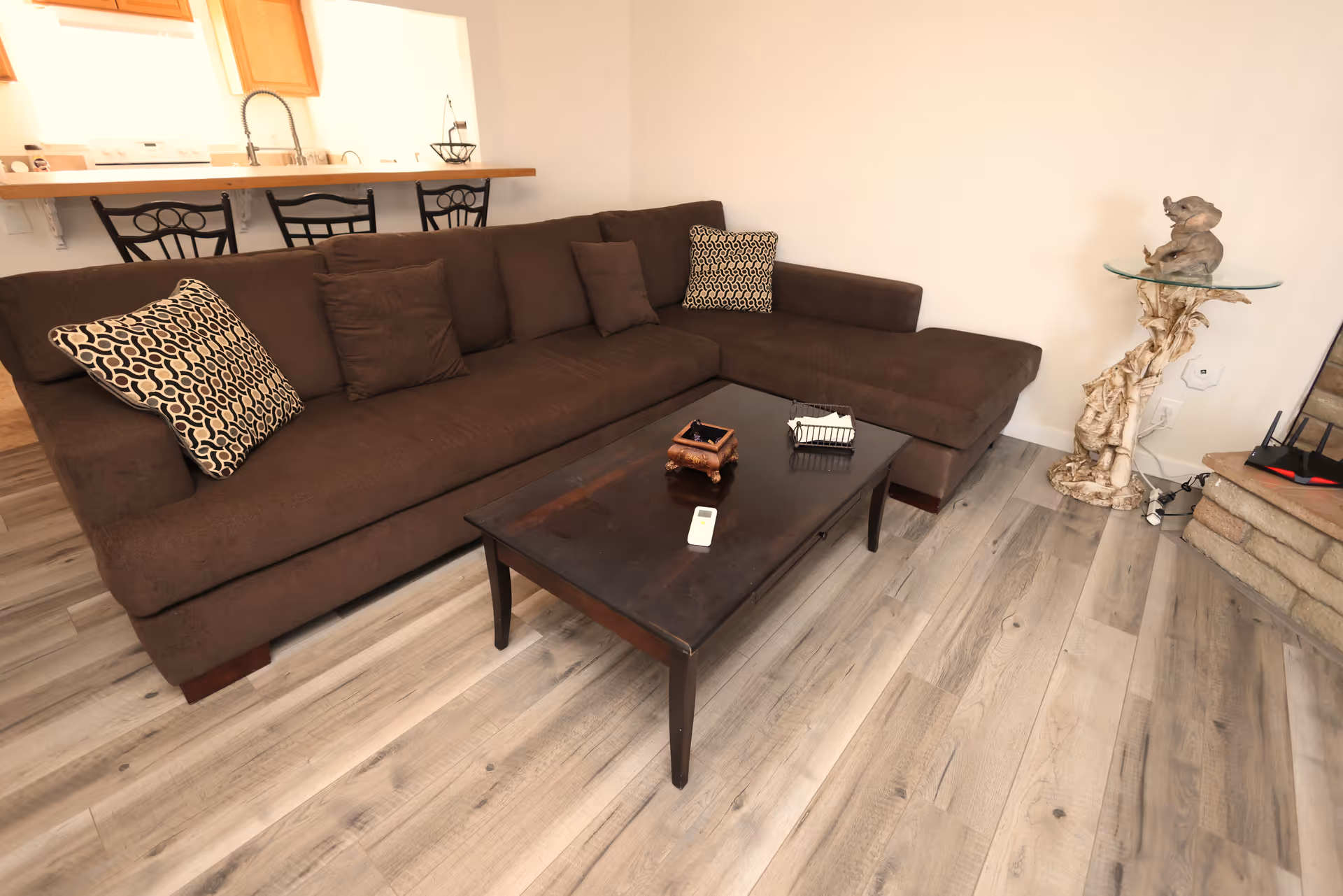 A cozy living room area with a large brown sectional sofa adorned with patterned and solid pillows. In front of the sofa is a dark wooden coffee table holding a small decorative box, a remote control, and a small basket. Behind the sofa, there is a kitchen counter with three black metal bar stools. To the right, there is a unique glass side table with a sculpted base and a small elephant figurine on top. The room has light wood flooring and white walls.