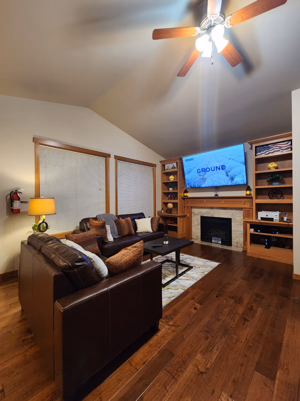 A cozy living room with wooden floors, two brown leather sofas with multiple cushions, a black coffee table on a patterned rug, a fireplace with a mounted TV above displaying the word 'GROUND', built-in wooden shelves on either side of the fireplace decorated with various items, and a ceiling fan with lights.