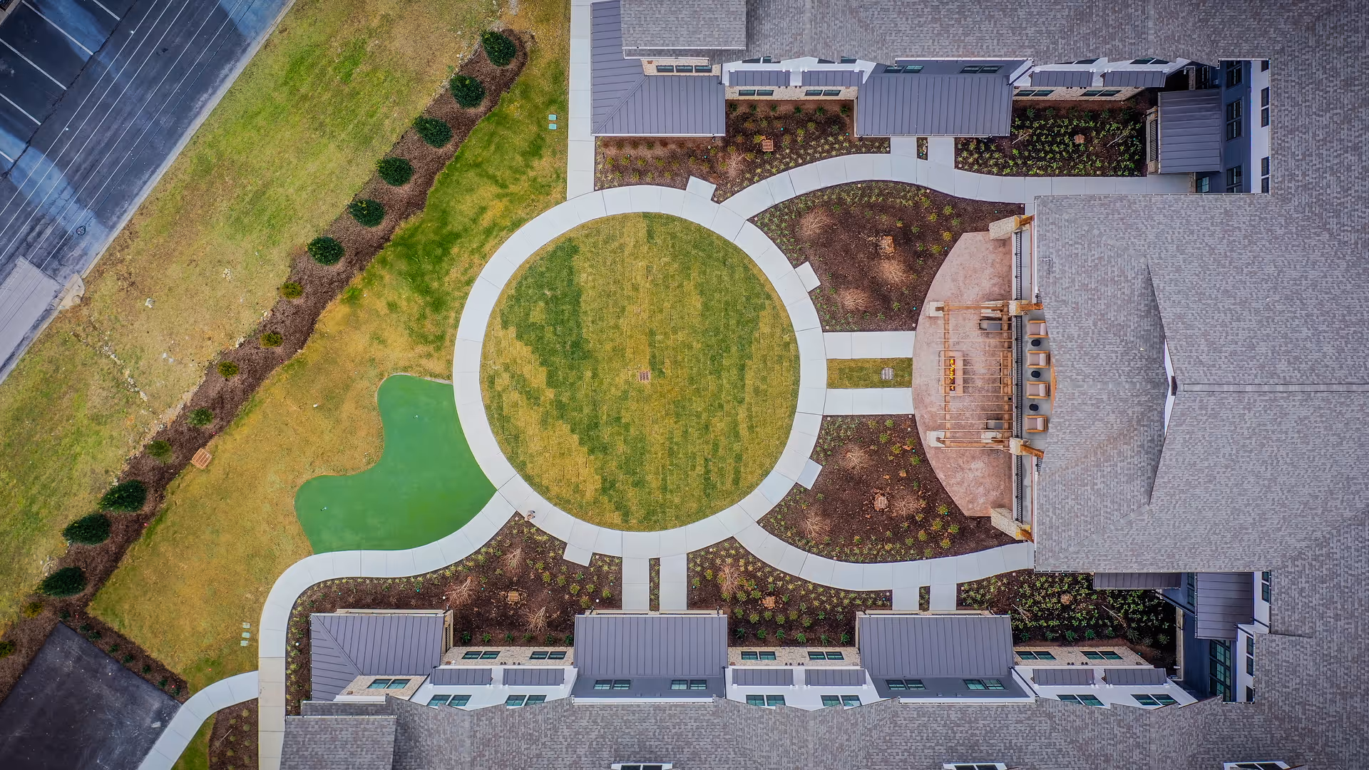 Aerial view of a senior living facility courtyard with a circular grassy area surrounded by a curved walkway, landscaped garden beds, and multiple building wings with gray roofs.