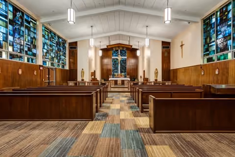 Interior view of a chapel with wooden pews arranged in rows facing an altar. The walls are adorned with stained glass windows on the left side and religious statues and a crucifix on the right. The ceiling has hanging lights, and the floor is covered with a patterned carpet.
