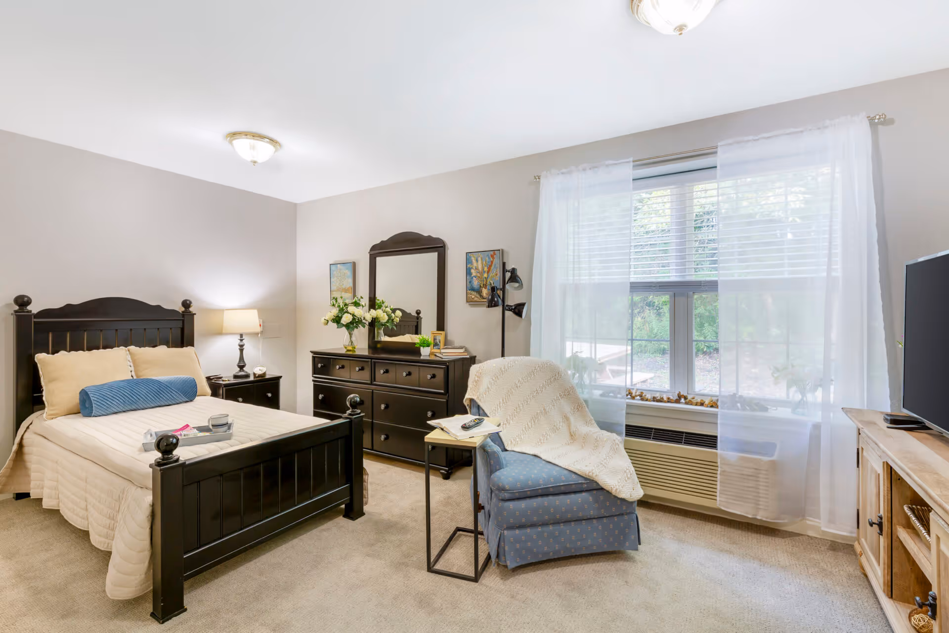 A cozy bedroom with a black wooden bed frame, beige bedding, and a blue cylindrical pillow. There is a black dresser with a mirror above it, decorated with flowers and framed pictures. A blue armchair with a white knitted throw blanket is positioned near a window with sheer white curtains. A small side table next to the chair holds a book and a remote control. A TV sits on a wooden stand opposite the bed. The room has beige carpet and neutral-colored walls.