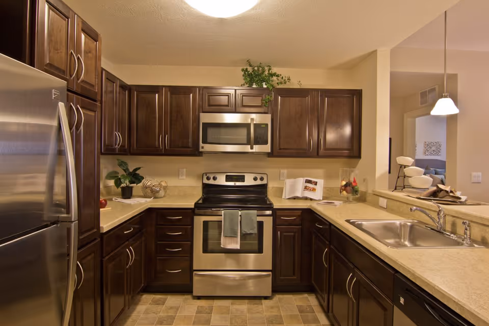 Modern kitchen with dark wooden cabinets, stainless steel refrigerator, oven, microwave, and dishwasher. The kitchen has beige countertops, a double sink, and a small plant and decorative items on the counter. There is a pendant light hanging above the counter that opens to a living area.
