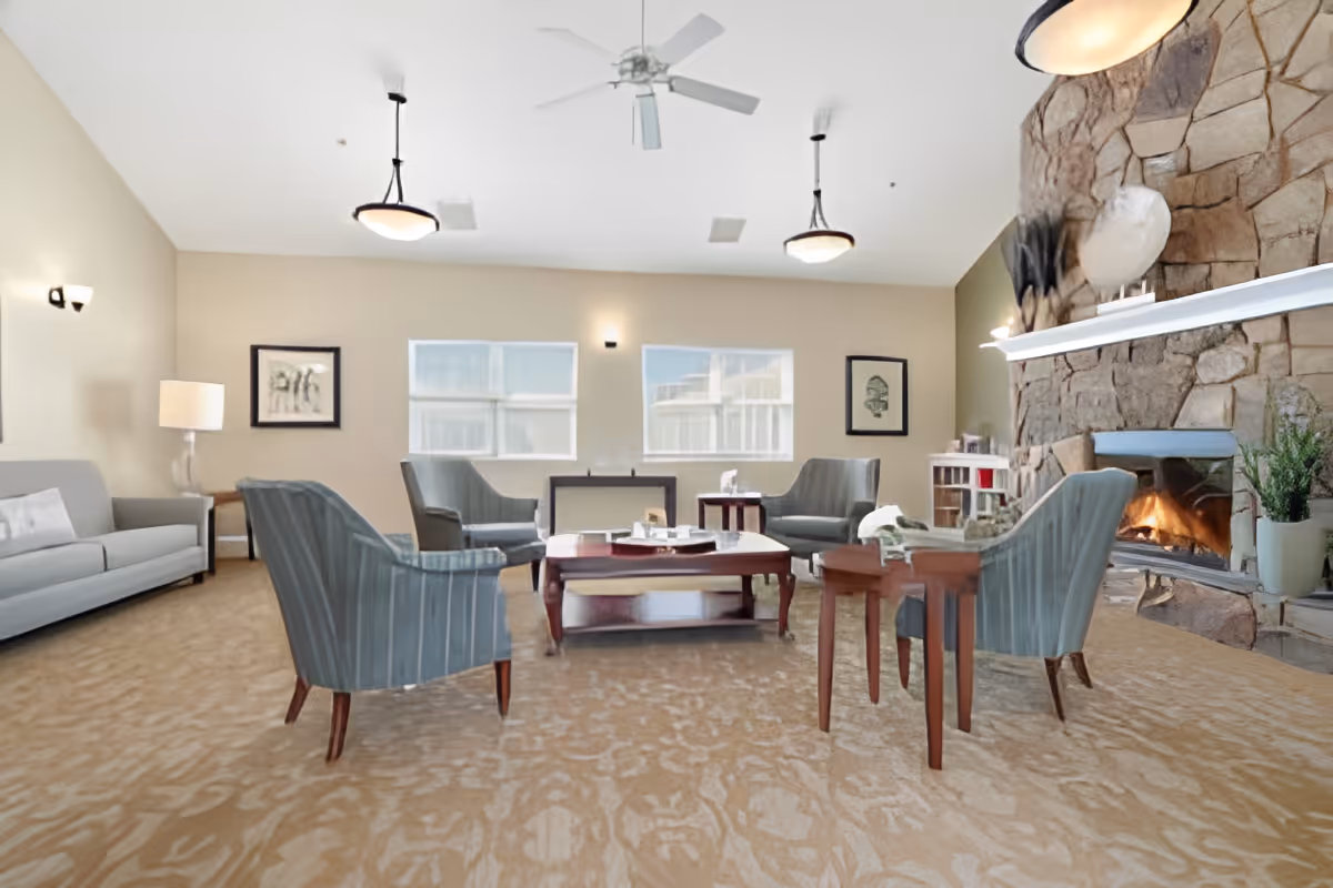 Spacious assisted living common room with armchairs and sofas arranged around a coffee table in front of a stone fireplace.