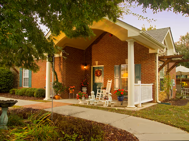 Front porch of a brick building with white columns and a green door decorated with a wreath. There are two white rocking chairs, potted plants, and small statues on the porch. A curved concrete walkway leads to the entrance, and trees and shrubs surround the area.