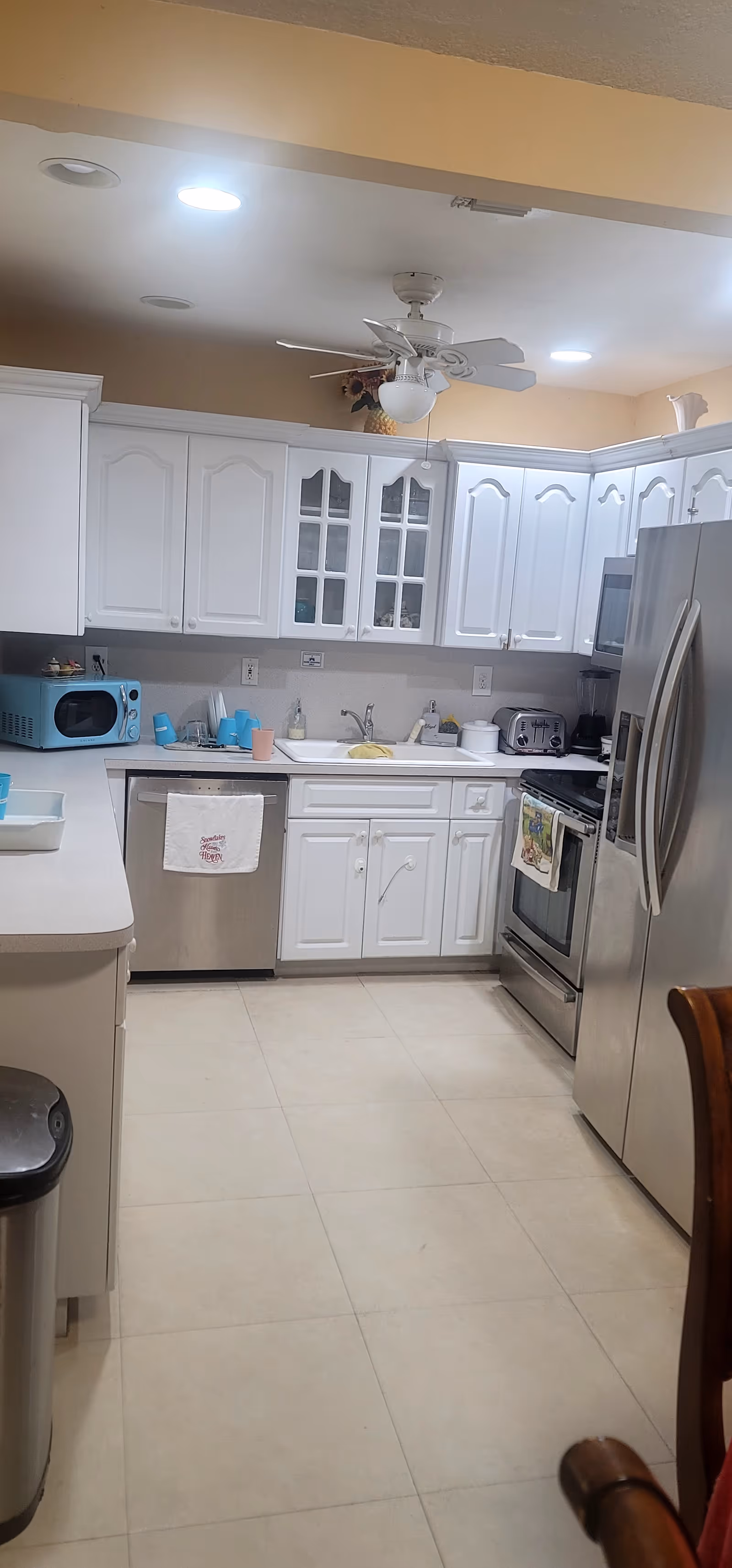A clean, modern kitchen with white cabinets and drawers, stainless steel refrigerator, oven, and dishwasher. There is a ceiling fan with a light fixture, a blue microwave on the counter, a toaster, and various kitchen items near the sink. The floor is tiled in a light color.