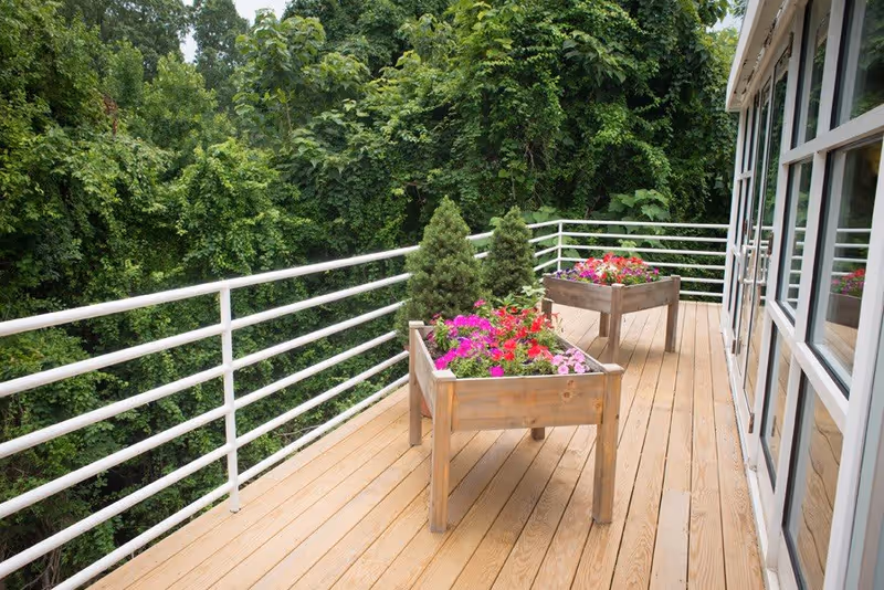Wooden deck with white metal railing, featuring two raised wooden planters filled with colorful flowers and small evergreen trees, adjacent to a building with large windows and surrounded by dense green foliage.
