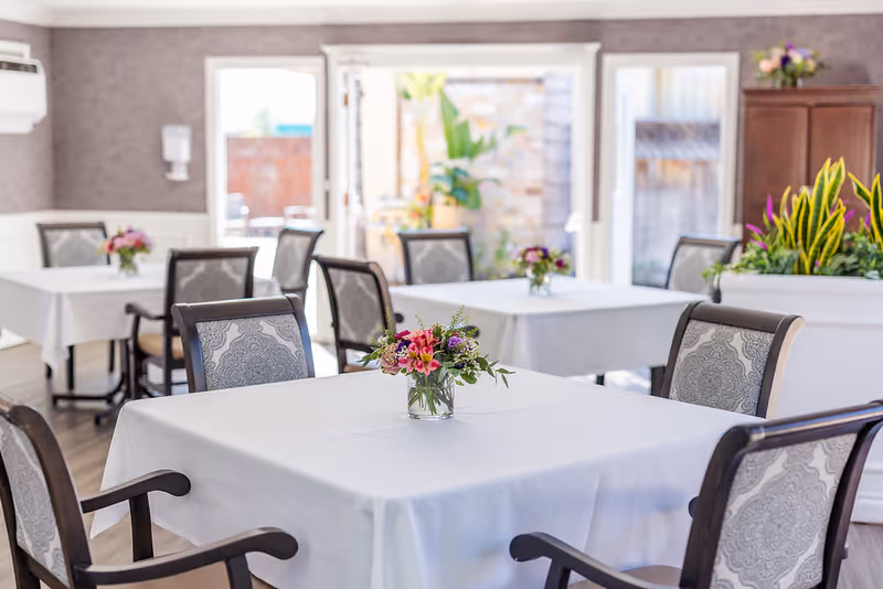 A bright dining room with tables covered in white tablecloths, each adorned with a small vase of colorful flowers. The room has cushioned chairs with patterned upholstery, large windows and glass doors letting in natural light, and a planter with green plants in the background.