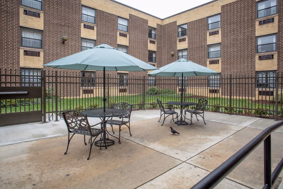 Fenced courtyard with patio tables and umbrellas, metal chairs, and a pigeon in front of a multi-story brick building.