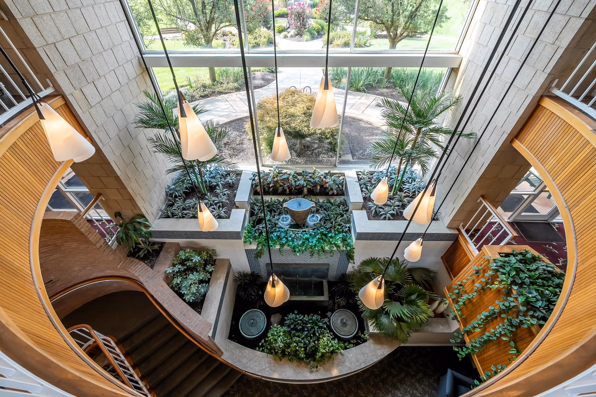 View from above of an indoor garden area with various green plants and a small water feature, surrounded by a curved staircase and large windows letting in natural light.