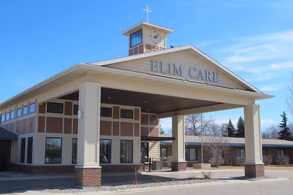Exterior view of the Fargo Elim Health Care Center building showing the entrance with a large covered drop-off area supported by columns. The building has a clock tower with a cross on top and the words 'ELIM CARE' displayed prominently above the entrance. The sky is clear and blue.