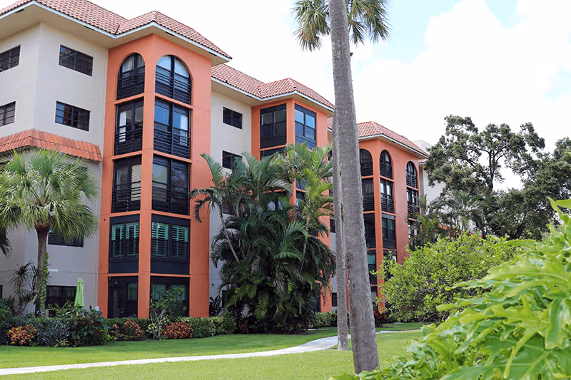 Exterior view of a multi-story Mediterranean-style senior living building with arched windows, palm trees, and landscaped lawn.