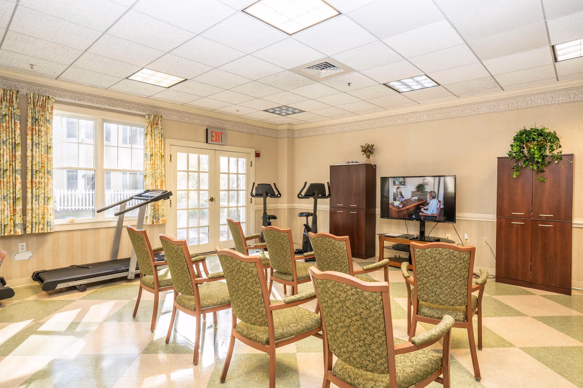 A bright room with several green upholstered chairs arranged in rows facing a flat-screen TV mounted on a stand. The room has exercise equipment including a treadmill and two stationary bikes near a set of French doors with an exit sign above. There are two wooden cabinets, one with a plant on top, and floral curtains on the windows letting in natural light.