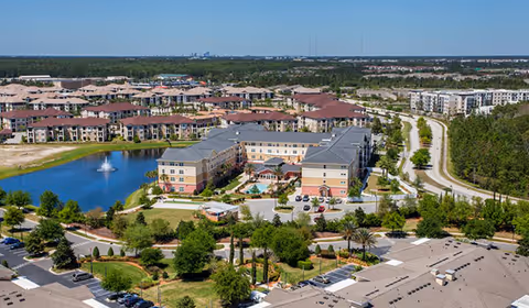 Aerial view of a senior living facility named Camellia At Deerwood, showing multiple buildings with red and gray roofs surrounding a pond with a fountain. The area is landscaped with trees and greenery, and there is a road curving around the property. In the background, a distant city skyline is visible under a clear blue sky.