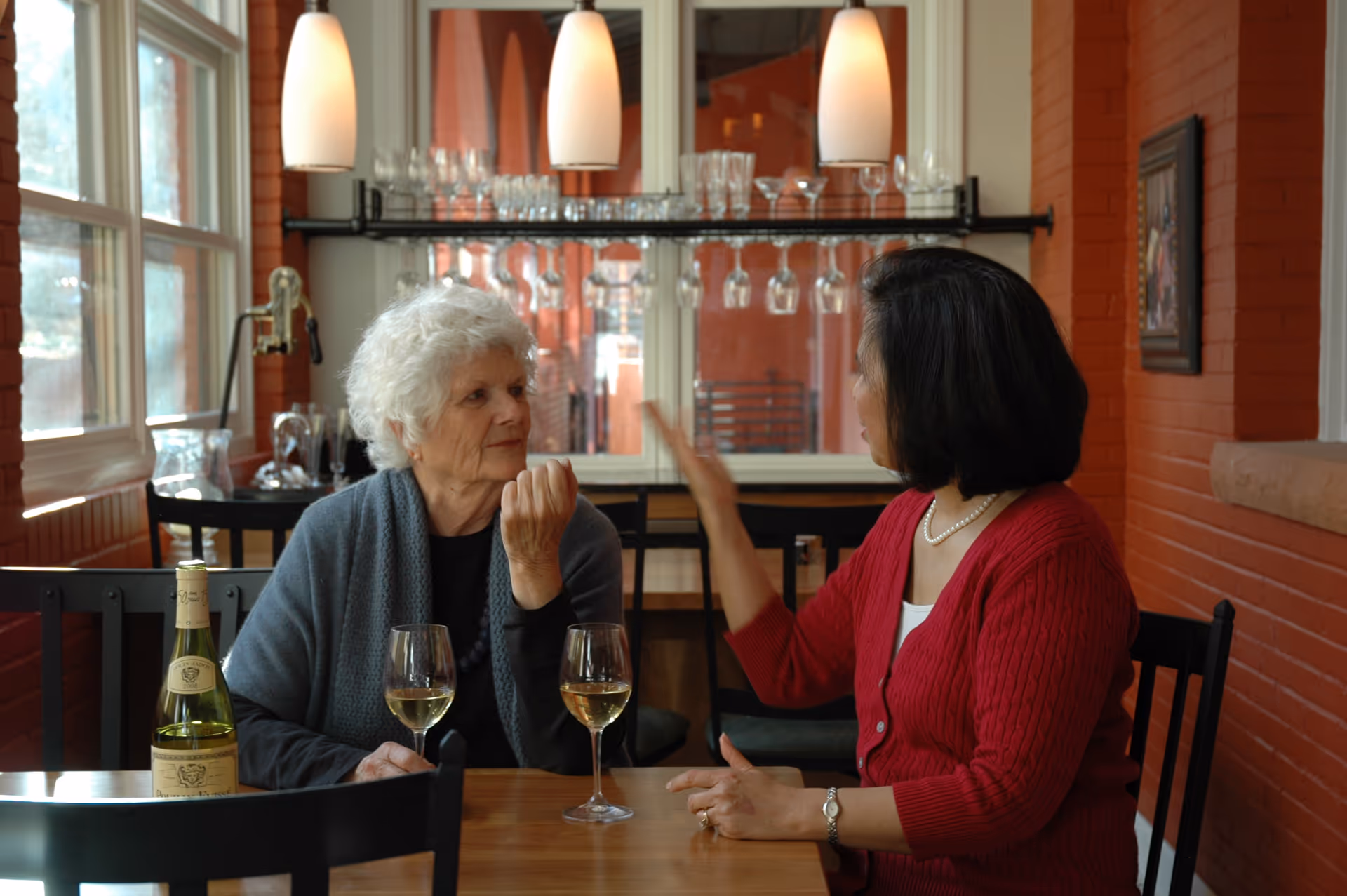 Two women sit at a table in a cozy dining area with wine glasses and a bottle of wine.