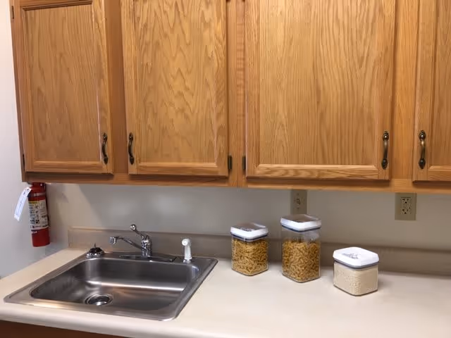 Kitchen countertop with a stainless steel sink and faucet, three clear containers with white lids holding pasta and rice, wooden cabinets above, and a fire extinguisher mounted on the wall to the left.