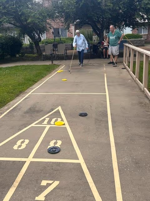 Two elderly people playing shuffleboard on an outdoor court at a senior living facility. The court is marked with numbers and lines, and there is greenery and trees surrounding the area. One person is actively playing while the other watches.